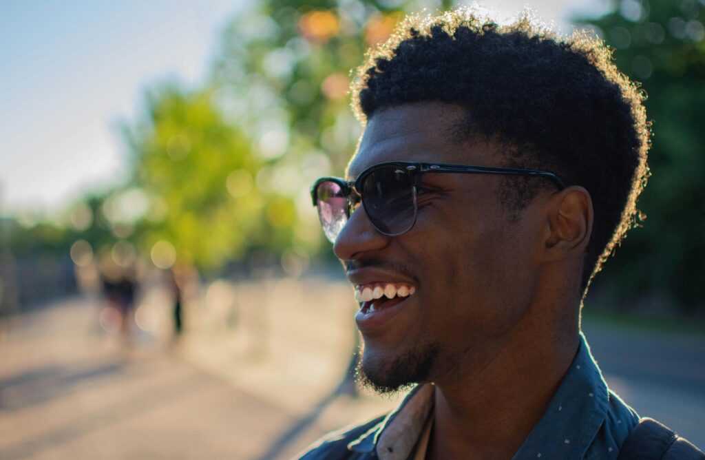 A smiling African American man wearing sunglasses, outdoors on a sunny day.